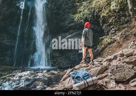 Femme avec sac à dos habillé de chapeau rouge et vêtements de randonnée actifs debout près de la cascade de la rivière de montagne et appréciant la puissance de la nature éclabousse. Banque D'Images
