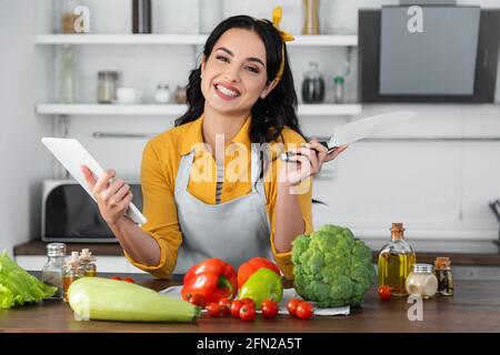 bonne femme tenant la tablette numérique tout en tenant le couteau près des légumes sur la table Banque D'Images