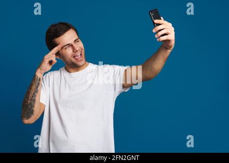 Un gars joyeux qui pointe les doigts sur son temple tout en prenant le selfie sur un téléphone portable isolé sur fond bleu Banque D'Images