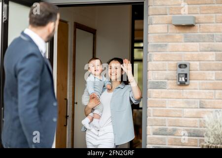 Un père caucasien agite Au revoir à sa famille avant d'aller travailler. Femme au foyer avec un nouveau-né restant devant la porte d'entrée et disant Au revoir à son mari. Style de vie familial. Banque D'Images