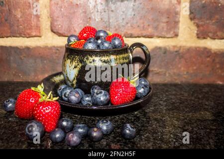 Encore-vie consistant d'une tasse noire remplie à déborder de fraises et de bleuets, avec la papillon plus entourant, contre un mur de brique. Banque D'Images