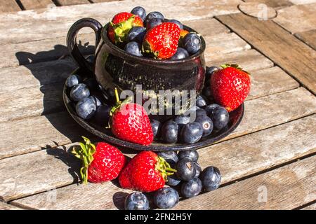 Une vie plate composée d'une tasse de thé noire remplie à déborder de fraises et de bleuets sur une surface en bois abîmé Banque D'Images