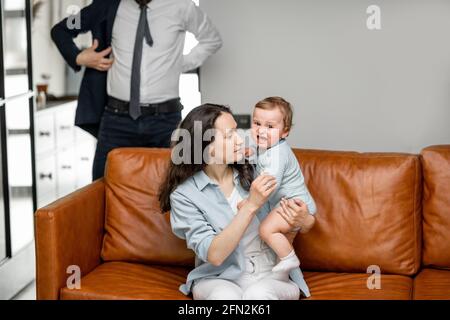 Une jeune maman assise avec son nouveau-né sur un canapé et prendre soin ou jouer avec un enfant pendant que le père et le mari vont travailler et dire Au revoir. Banque D'Images