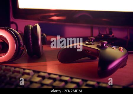 Vue de la configuration du bureau de jeu avec une manette de jeu noire, un casque, un clavier et un moniteur illuminés par des lumières rouges et violettes. Banque D'Images