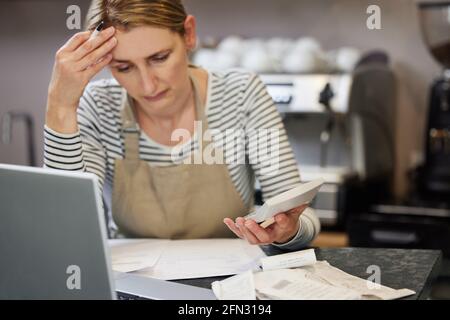 Femme inquiète propriétaire du café en difficulté financière Via des factures utilisant un ordinateur portable et une calculatrice Banque D'Images