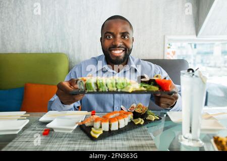 Un jeune homme d'affaires africain assis au bar à sushis, qui fait une démonstration à la grande assiette de l'appareil photo avec un ensemble de petits pains à sushis, se moque de l'expression du visage Banque D'Images