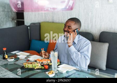 Un Africain est assis dans un restaurant, parlant au téléphone et mangeant des sushis japonais. Un homme d'affaires souriant mange des sushis avec des baguettes et parle avec un ami pendant le déjeuner Banque D'Images