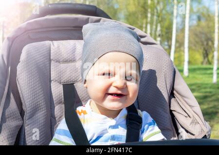 Adorable bébé caucasien heureux assis dans une poussette grise dans le parc. Jour d'été. L'enfant est attaché dans la poussette pendant la marche. Banque D'Images