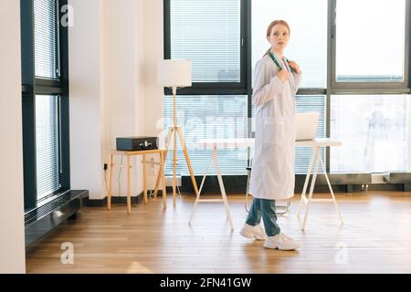 Portrait d'une femme médecin pendieuse en manteau blanc debout sur fond de fenêtre en journée ensoleillée dans le bureau de clinique médicale légère. Banque D'Images