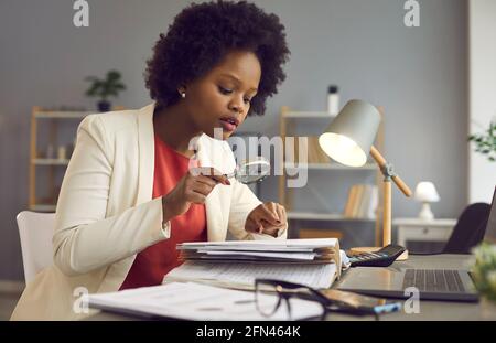 Une femme d'affaires afro-américaine concentrée lit et étudie des documents avec une loupe. Banque D'Images