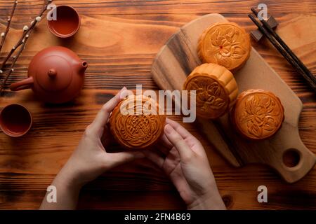 Vue de dessus des mains des femmes tenant un gâteau traditionnel de lune avec un plateau de thé et des gâteaux de lune sur une table rustique. Le caractère chinois sur le gâteau de lune représente 'cinq Banque D'Images