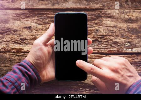 man tient un téléphone portable avec un écran noir vierge. lit les messages à l'écran du smartphone. sur fond de maquette en bois Banque D'Images