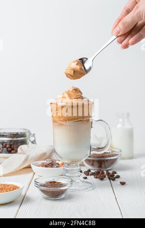 La main d'une femme met de la mousse dans un verre de café de Dalgona sur un fond blanc. Boisson au café coréen avec lait et glace. Banque D'Images
