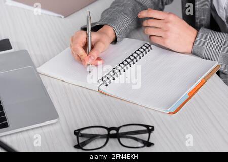 Vue rognée d'une femme d'affaires écrivant sur un carnet à proximité de lunettes et ordinateur portable Banque D'Images