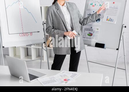 Vue rognée d'une femme d'affaires avec un smartphone pointant vers des graphiques tableau à côté de l'ordinateur portable Banque D'Images