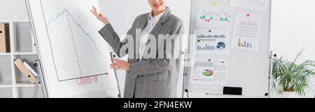 Vue rognée d'une femme d'affaires souriante pointant sur un paper-board avec un lettrage Covid-2019 et une bannière Banque D'Images