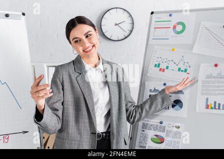 Femme d'affaires souriante dans des écouteurs pointant vers le tableau de conférence pendant la réunion en ligne Banque D'Images