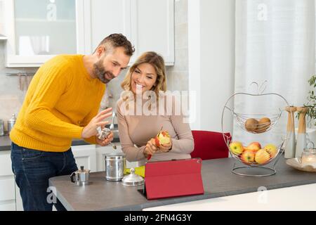 Bon couple qui fait un appel vidéo sur une tablette. Des jeunes mariés en relation avec des parents pendant le petit déjeuner. Un couple italien prépare des fruits et du café. Banque D'Images