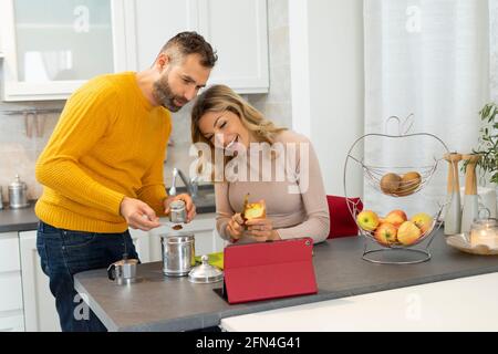 Bon couple qui fait un appel vidéo sur une tablette. Des jeunes mariés en relation avec des parents pendant le petit déjeuner. Un couple italien prépare des fruits et du café. Banque D'Images