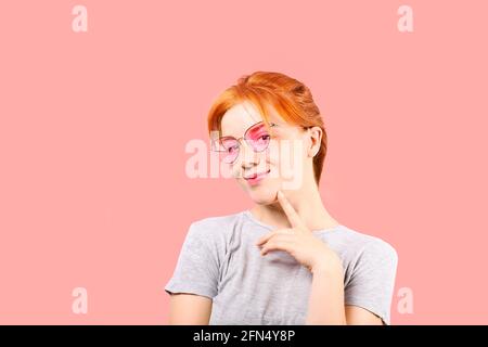 Jeune femme attrayante avec de longs cheveux rouges, portant des lunettes de soleil roses pour les yeux de chat, montrant un langage corporel dégoûtant, touchant le visage. Belle femme rouge Banque D'Images