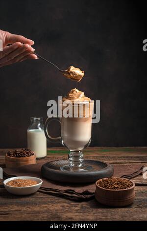 Une main de femme avec une cuillère et de la mousse fouettée et un verre de café de Dalgona sur fond marron. Boisson revigorante coréenne. Vue latérale, vue verticale. Banque D'Images