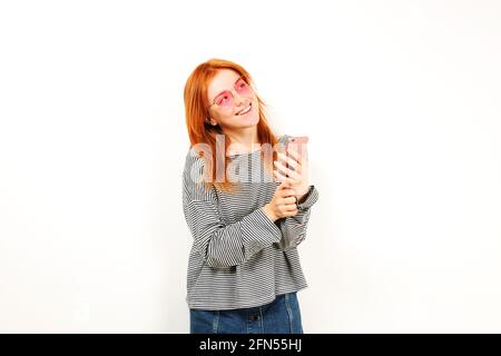 Jeune femme élégante à tête rouge naturelle souriant sur l'écran du smartphone, utilisant un téléphone portable, portant une chemise grise et des lunettes pour chats. Espace de copie blanc Banque D'Images
