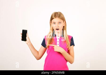 Portrait d'une jeune fille d'âge décontracté avec de longs cheveux blonds naturels, index pointant vers un téléphone cellulaire à écran vierge. Émerveillement jeune femme tenant smartphon Banque D'Images