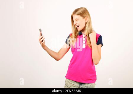 Heureusement belle jeune femme heureuse avec des cheveux longs blonds naturels célébrant sa victoire, criant et riant, regardant son écran de smartphone. Adolescent Banque D'Images