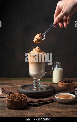 La main d'une femme met de la mousse de café dans un verre de café de Dalgona sur un fond marron. Vue verticale. Banque D'Images