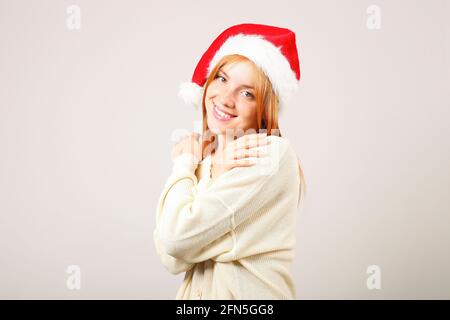 Portrait en gros plan d'une belle jeune femme à tête rouge portant un chapeau et un chandail blanc du Père Noël avec une expression faciale satisfaite. Mignonne célébrant Banque D'Images