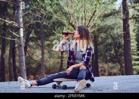 Une jeune femme assise sur un skateboard buvant de la bière. Banque D'Images