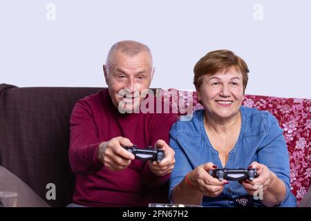 Vue de face de personnes âgées grises excitées, couple d'hommes et de femmes jouant à un jeu vidéo sur une console, assis sur un canapé coloré dans la vie Banque D'Images