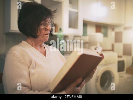 Femme âgée assise dans la buanderie et livre de lecture Banque D'Images