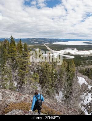 Homme debout sur le bord d'une falaise en veste bleue avec un vaste paysage du nord du Canada en dessous dans la forêt boréale pendant le printemps. Banque D'Images