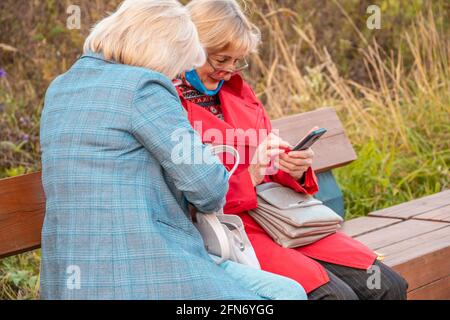 Kazan, Russie - 03 octobre 2020 : deux femmes en âge de prendre leur retraite assises sur un banc dans un parc municipal regardent l'écran du smartphone à travers leurs lunettes Banque D'Images