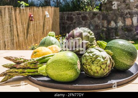 Concept de repas de légumes sains : une assiette d'artichauts verts et biologiques frais, d'asperges, de courgettes et de citrons mûrs jaunes sur une table dans le jardin Banque D'Images