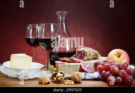 Salami, fromage, noix, raisins et vin rouge. Deux verres avec carafe sur table en bois et arrière-plan rouge avec espace pour le texte. Gros plan, vie fixe. Banque D'Images