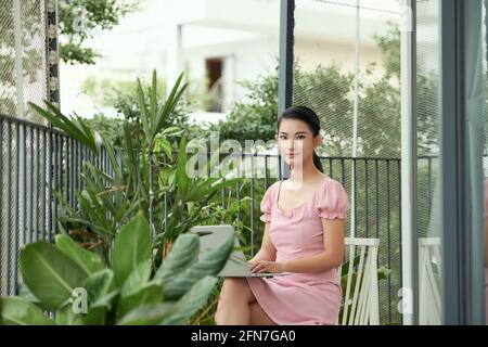 une femme heureuse et confiante utilisant un ordinateur portable tout en étant assise sur le balcon et en travaillant de chez elle. Banque D'Images