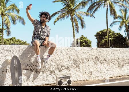 Jeune latino s'amusant à prendre un selfie avec un smartphone mobile tout en écoutant de la musique avec un casque et une boombox pendant les vacances d'été Banque D'Images