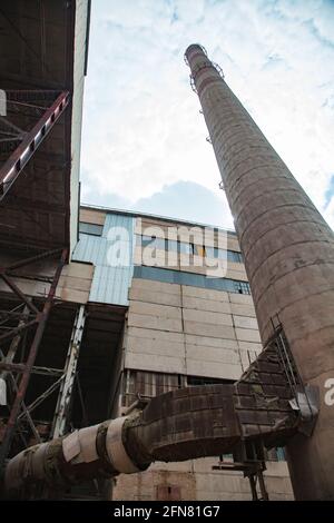 Temirtau, Kazakhstan. Une usine de ciment soviétique dépassée, un point de vue abstrait. Cheminée en béton avec tube rouillé et bâtiment industriel. Ciel bleu avec nuages Banque D'Images