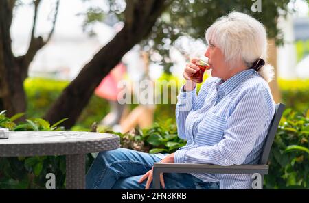 Femme sirotant un verre de thé chaud à l'extérieur patio car elle passe une journée de détente à regarder les gens marcher par derrière les plantes tropicales Banque D'Images