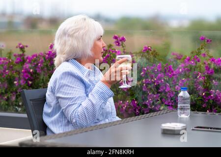 Belle femme d'âge moyen se détendant sur un patio ou un balcon extérieur au printemps avec un verre de vin blanc froid au-dessus de bougainville coloré Banque D'Images