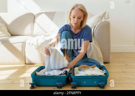 Femme mûre et attentionnés des années 60 qui a fait ses valises pour voyager tout en étant assise sur un parquet à la maison. Femme de direction se prépare pour les vacances d'été. Concept de voyage à la retraite Banque D'Images