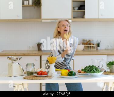 Femme sénieuse et insouciante en tablier utilisant une fourchette en bois comme microphone et chant, s'amusant tout en cuisinant des aliments sains dans la cuisine moderne et légère à la maison. Bonne femme mature qui profite de la vie à la retraite Banque D'Images
