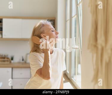 Vue latérale de belle femme sénior des années 60 grand-mère en tenue décontractée peignant les cheveux tout en se tenant devant la fenêtre dans la cuisine moderne, accent sélectif sur la femme mature. Concept de mode de vie des personnes âgées Banque D'Images