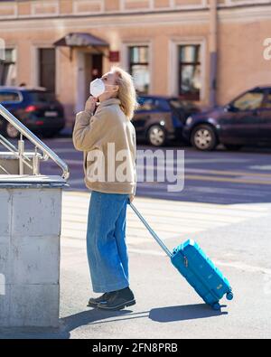 Femme voyageur senior à la mode portant un respirateur ou un masque facial debout sur une rue ensoleillée avec une valise bleue et regardant vers le haut. Une dame âgée se perd dans la ville pendant son voyage pendant l'épidémie de covid19 Banque D'Images