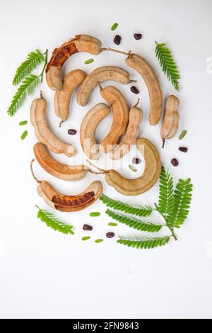 Gousses de type Tamarind  haricot remplies de graines entourées d'une pulpe fibreuse disposée dans un fond blanc texturé avec sa feuille verte fraîche. Banque D'Images