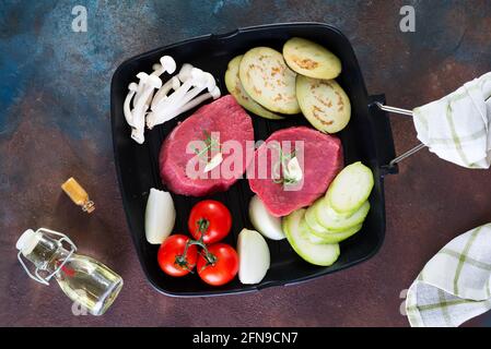 Steak de bœuf cru sur l'os avec des légumes frais dans une casserole sur fond de pierre sombre, vue du dessus Banque D'Images