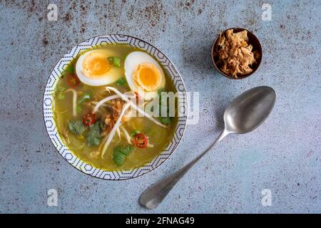 Un seul bol de Soto Ayam (traduction : soupe de poulet indonésienne) avec un œuf dur, un oignon frit et de la coriandre, d'en haut sur un backgroun bleu texturé Banque D'Images