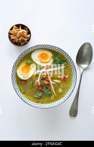 Un seul bol de Soto Ayam (traduction : soupe de poulet indonésienne) avec un œuf dur, un oignon frit et de la coriandre, d'en haut sur fond blanc, copie Banque D'Images